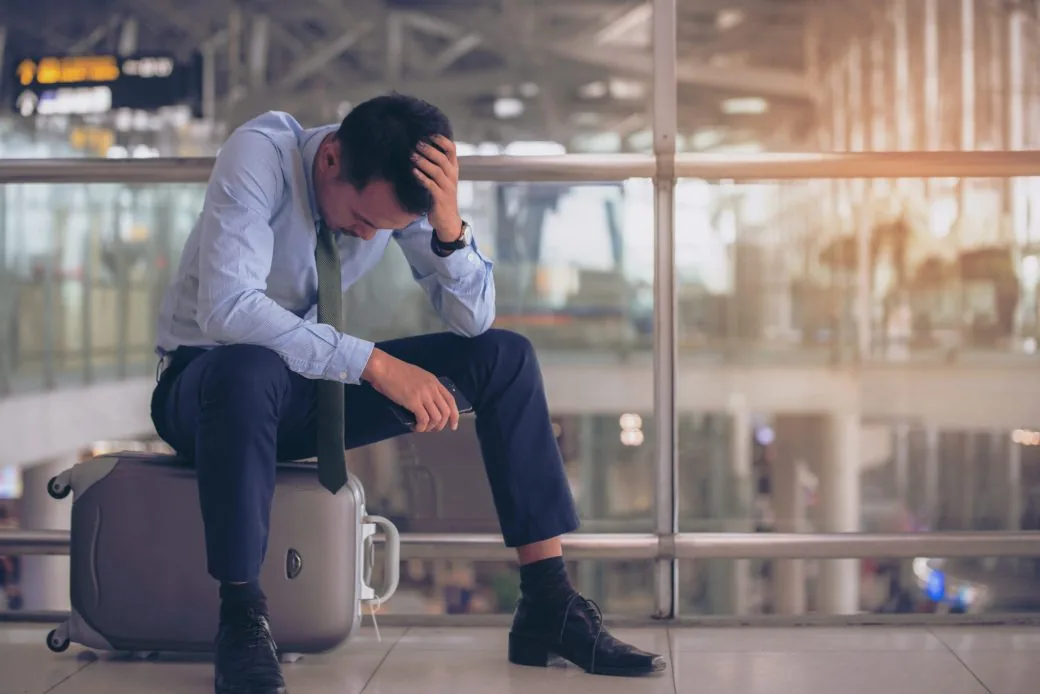 A tired traveller sitting on a suitcase with his head in his hand at an airport terminal.