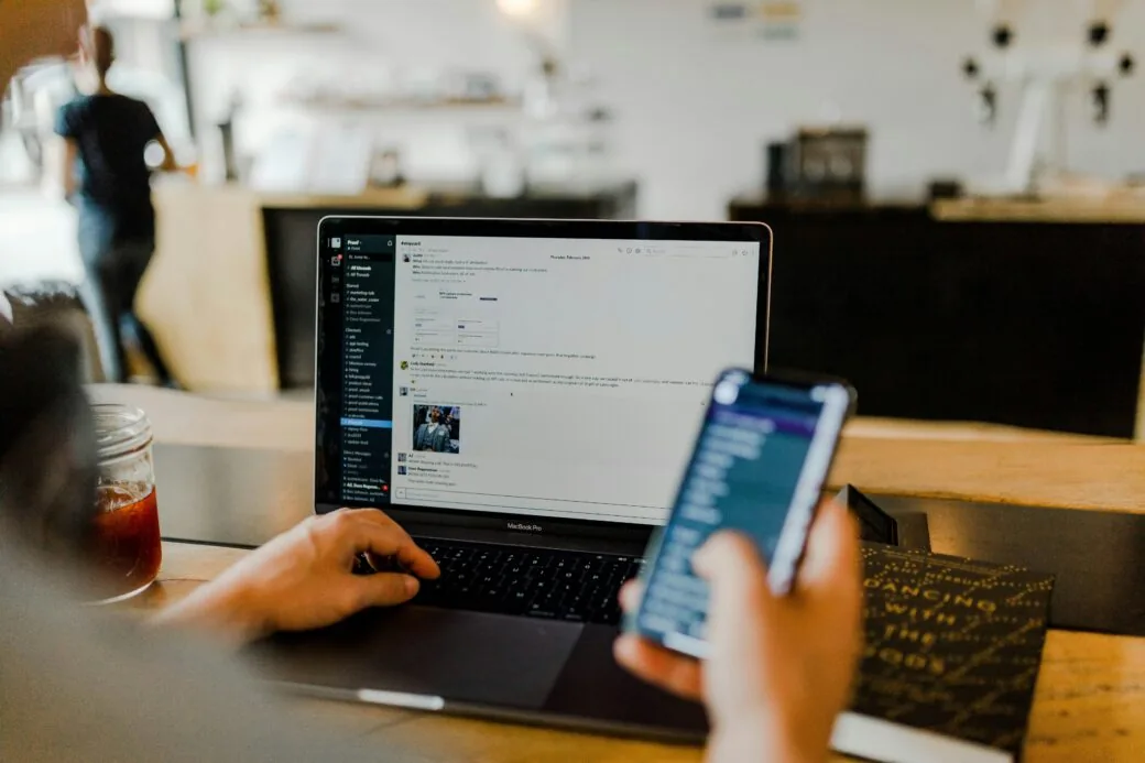 A person typing on a laptop while holding a smartphone at a cafe table with a drink beside them.