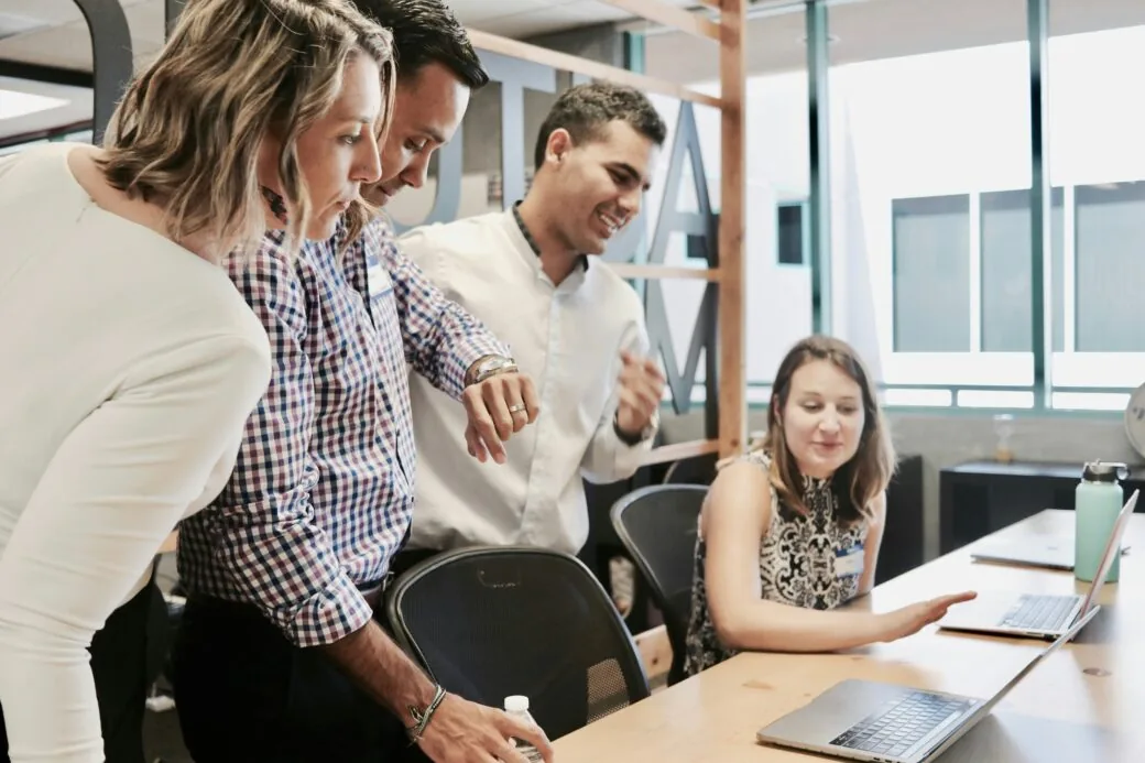 Four colleagues gathered around a laptop, smiling as they look at the screen together in an office.
