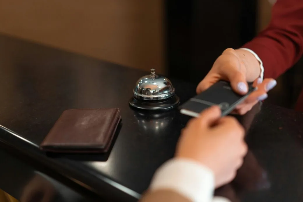 Hands exchange a room key card over a hotel reception desk with a bell and wallet nearby.