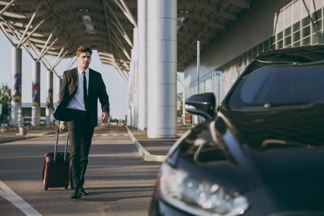 A businessman pulling a suitcase towards a car under an airport pick-up canopy.