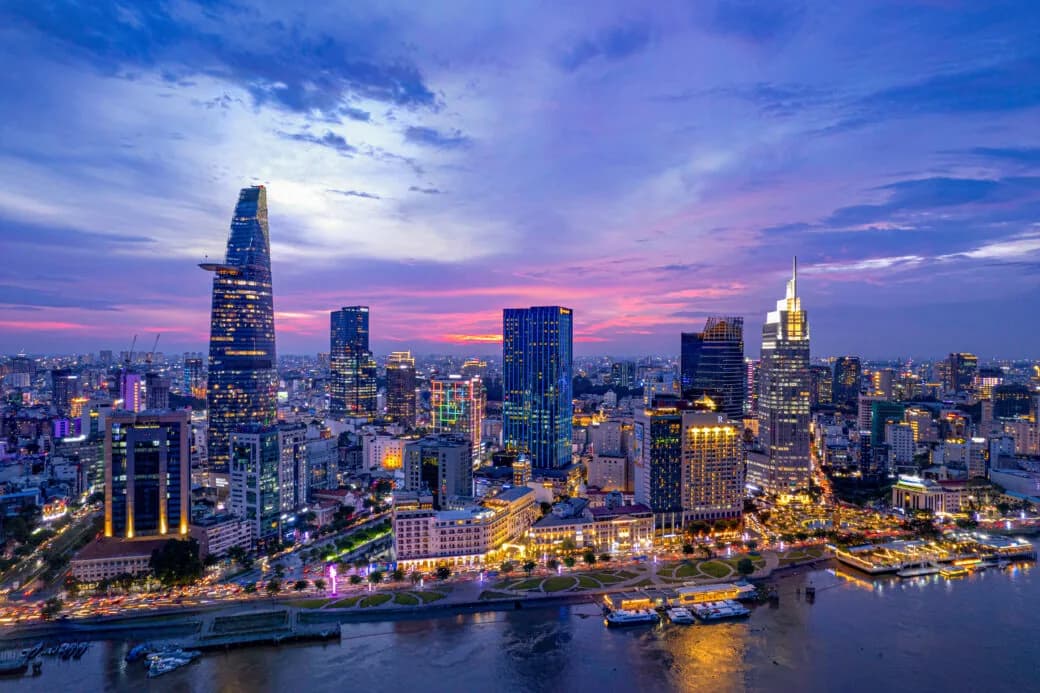 A city skyline at dusk with illuminated skyscrapers along a riverside promenade and boats on the water.