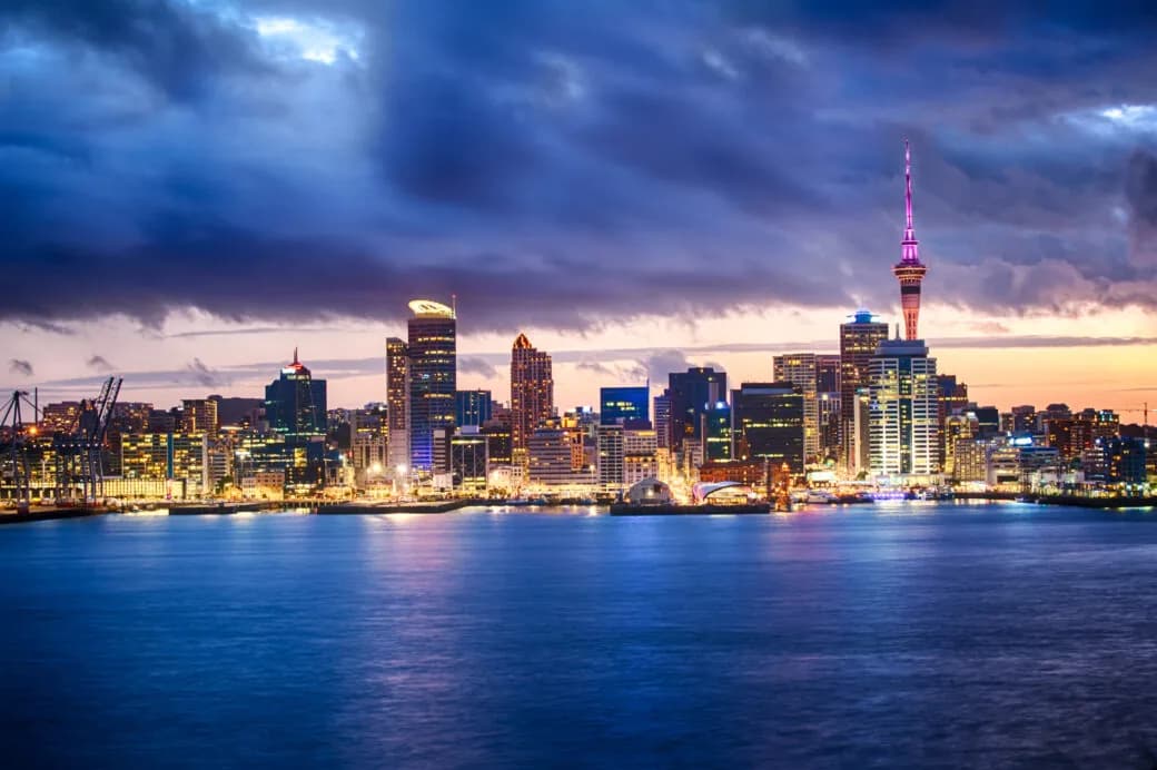 A city skyline at dusk with a tall illuminated tower rising above the waterfront under a cloudy sky.